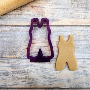 May include: A purple plastic cookie cutter shaped like a one-piece swimsuit. A brown sugar cookie in the shape of a one-piece swimsuit is next to the cutter.
