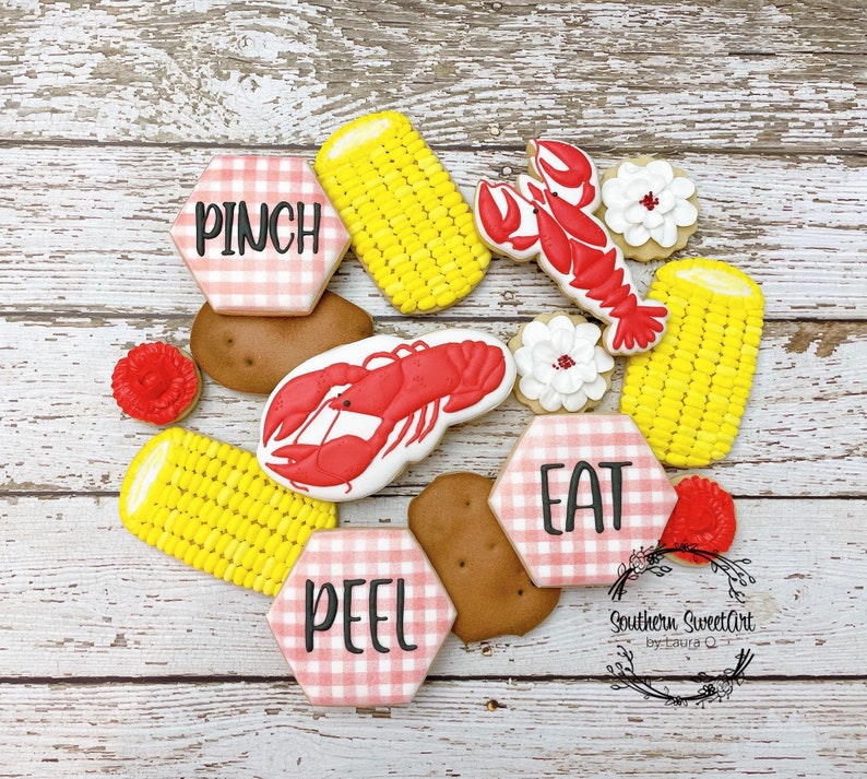 May include: A set of nine decorated sugar cookies with a red, white, and blue gingham pattern. The cookies feature images of corn on the cob, lobsters, and the words "Pinch", "Peel", and "Eat".