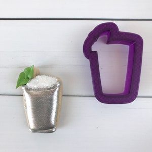 May include: A silver cookie decorated to look like a cocktail glass with a green leaf and white sugar on top. A purple plastic cookie cutter in the shape of a cocktail glass is also shown.