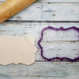 Pode incluir: Um cortador de biscoitos de plástico roxo em forma de retângulo com cantos arredondados e bordas decorativas. Um recorte de massa de biscoito branco simples está ao lado do cortador.