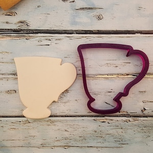 May include: A close-up of two cookie cutters, one is a light beige color and the other is a dark purple color. Both cookie cutters are shaped like a teacup.