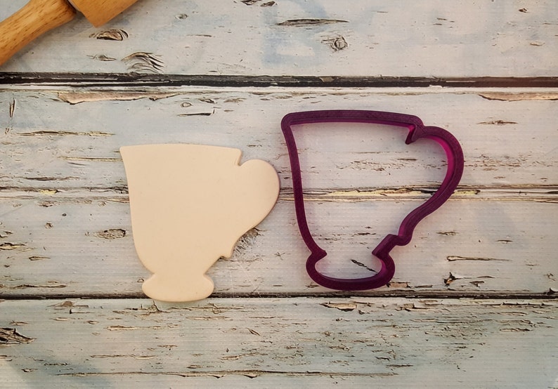 May include: A close-up of two cookie cutters, one is a light beige color and the other is a dark purple color. Both cookie cutters are shaped like a teacup.