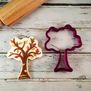 May include: A cookie cutter and a decorated cookie in the shape of a tree with brown branches and colorful leaves. The cookie cutter is purple and the cookie is on a white wooden surface.