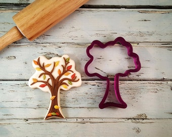 May include: A cookie cutter and a decorated cookie in the shape of a tree with brown branches and colorful leaves. The cookie cutter is purple and the cookie is on a white wooden surface.