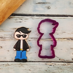 May include: A decorated cookie in the shape of a cartoon man wearing sunglasses, a black jacket, blue jeans, and black shoes. Next to it is a purple plastic cookie cutter in the same shape. A wooden rolling pin is in the background.