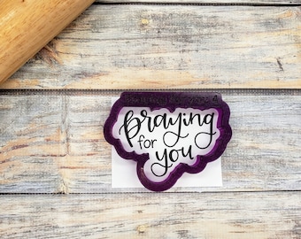 Op de afbeelding: Paarse plastic koekjesvormer in de vorm van een spreekballon met de tekst "Praying for you" in zwarte letters.