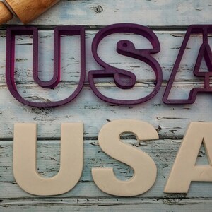 May include: Purple plastic cookie cutters in the shape of the letters U, S, and A.  Below the cutters are white cookie dough letters that spell out USA.