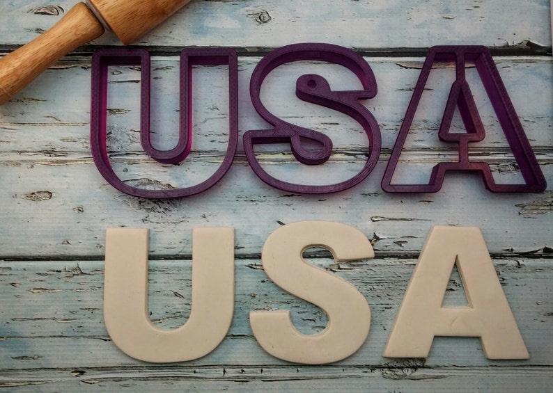 May include: Purple plastic cookie cutters in the shape of the letters U, S, and A.  Below the cutters are white cookie dough letters that spell out USA.