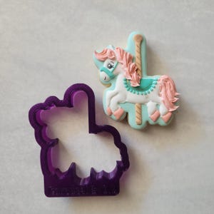 May include: A carousel horse cookie decorated with white icing, pink mane and tail, and a teal saddle. A purple plastic cookie cutter in the shape of the carousel horse is next to the cookie.