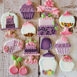 May include: A collection of 13 purple and pink decorated sugar cookies. The cookies are shaped like cupcakes, jars, cherries, and cakes. The cookies are decorated with flowers, hearts, and sprinkles. The cookies are arranged on a white wooden surface.