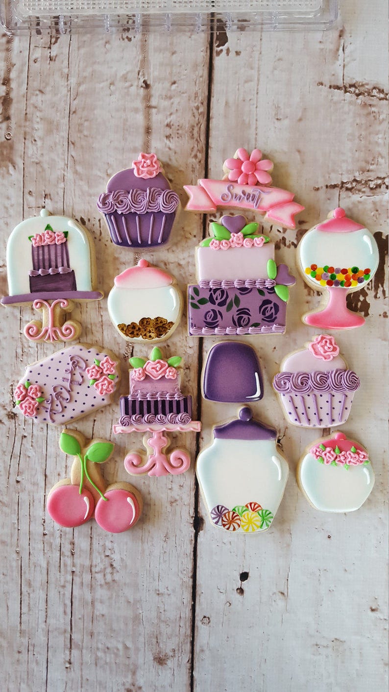 May include: A collection of 13 purple and pink decorated sugar cookies. The cookies are shaped like cupcakes, jars, cherries, and cakes. The cookies are decorated with flowers, hearts, and sprinkles. The cookies are arranged on a white wooden surface.