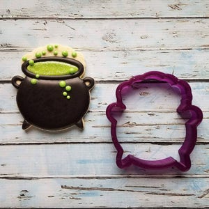 May include: A black and green cookie decorated to look like a cauldron with green bubbling liquid. A purple plastic cookie cutter in the shape of a cauldron is next to the cookie.