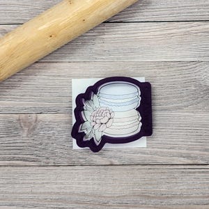 May include: Purple plastic cookie cutter shaped like a stack of macarons with a floral design. The macarons are light blue, pink, and white.
