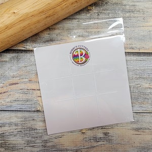 May include: A white plastic stencil with nine square cutouts. The stencil is in a clear plastic bag. The bag has a logo that says "Bobbi's Cutters" with a colorful letter B in the center.