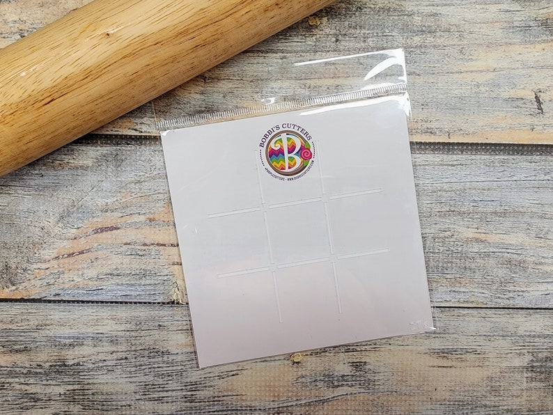 May include: A white plastic stencil with nine square cutouts. The stencil is in a clear plastic bag. The bag has a logo that says "Bobbi's Cutters" with a colorful letter B in the center.