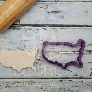 May include: A close-up of a purple plastic cookie cutter shaped like the United States of America. A beige piece of cookie dough is also in the image, shaped like the United States of America. A wooden rolling pin is in the background.