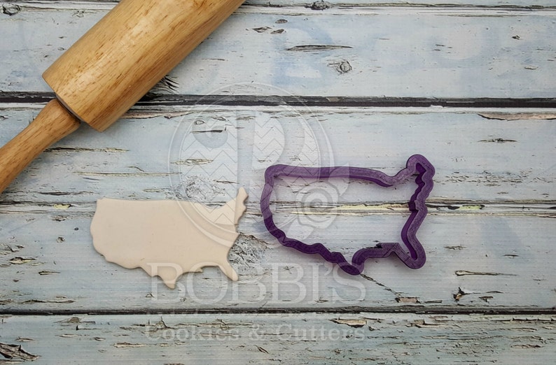 May include: A close-up of a purple plastic cookie cutter shaped like the United States of America. A beige piece of cookie dough is also in the image, shaped like the United States of America. A wooden rolling pin is in the background.