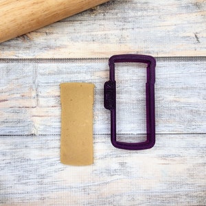 May include: Purple plastic cookie cutter shaped like a can. A piece of brown cookie dough is next to the cutter.