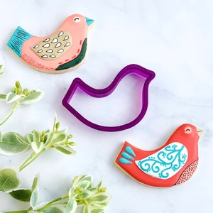 May include: Two decorated bird-shaped cookies and a purple plastic cookie cutter in the shape of a bird. The cookies are decorated with red, pink, blue, and white icing.