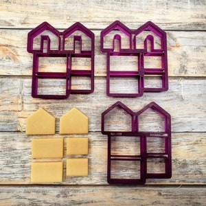Pode incluir: Quatro cortadores de biscoitos de plástico roxo em forma de casas com várias janelas e portas. Cinco pedaços de massa de biscoito bege estão dispostos em uma pilha abaixo dos cortadores.
