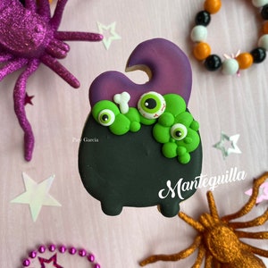 May include: A black and green Halloween cookie decorated with a purple witch's hat, googly eyes, and a bone. The cookie is decorated with the text "Mantequilla".