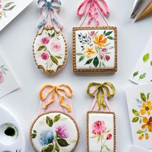 May include: Five decorated sugar cookies with floral designs. Each cookie is shaped like a picture frame with a bow at the top. The cookies are painted with watercolor-like designs of flowers and leaves in shades of pink, yellow, blue, and green.