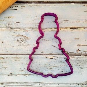May include: A purple plastic cookie cutter shaped like a Christmas tree. The cutter is on a white wooden surface.