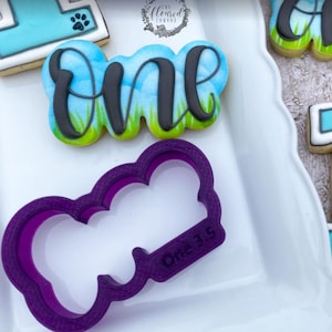 May include: A decorated sugar cookie with the word "one" in black script, set against a blue sky and green grass background. A purple cookie cutter with the text "One 3.5" is also present.