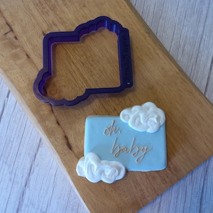 May include: A blue cookie decorated with white frosting clouds and the text "oh, baby" in gold. A purple plastic cookie cutter shaped like a rectangle with rounded corners is also shown.