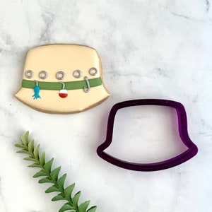 May include: A beige, fisherman's hat-shaped cookie decorated with green and silver accents, and fishing-themed details. A purple cookie cutter in the shape of the hat sits next to the cookie. The cookie has a brown border.