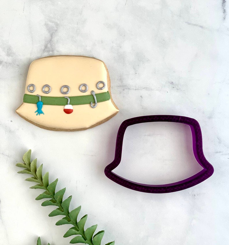 May include: A beige, fisherman's hat-shaped cookie decorated with green and silver accents, and fishing-themed details. A purple cookie cutter in the shape of the hat sits next to the cookie. The cookie has a brown border.