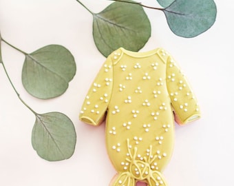 May include: A yellow sugar cookie decorated with white icing dots in the shape of a baby onesie. The cookie is on a white background with green eucalyptus leaves.