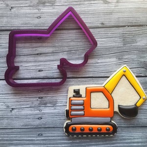 May include: A yellow and orange cookie decorated to look like an excavator. The cookie is on a wooden surface next to a purple plastic cookie cutter in the shape of a house.