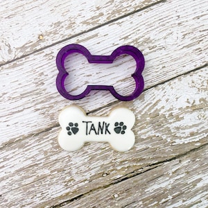 Peut inclure: Emporte-pièce en forme d'os de chien en plastique violet avec un biscuit en forme d'os blanc décoré d'empreintes de pattes noires et du mot "TANK" en glaçage noir.