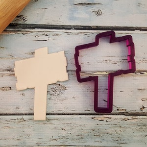 May include: A wooden sign cookie cutter with a purple plastic outline. The cookie cutter is on a white wooden surface.