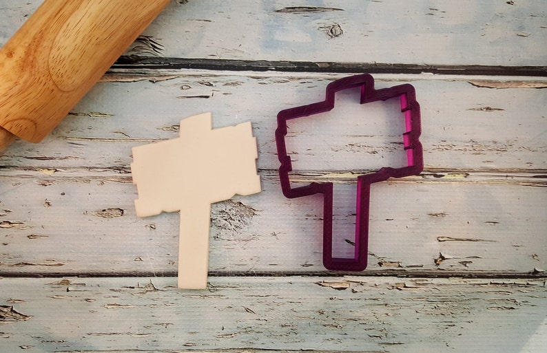 May include: A wooden sign cookie cutter with a purple plastic outline. The cookie cutter is on a white wooden surface.
