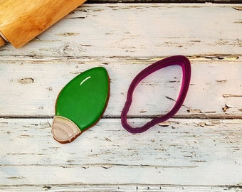 May include: A green Christmas light-shaped cookie with a brown outline and a light tan base, next to a purple cookie cutter. A wooden rolling pin rests in the upper left corner. The objects are on a white, distressed wooden surface.