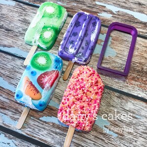 May include: Four different popsicle-shaped cakes decorated with various toppings, including kiwi, strawberries, and sprinkles. The cakes are on a wooden surface with a purple popsicle mould.