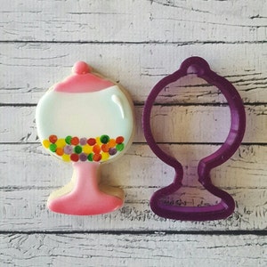 May include: A pink and white cookie cutter shaped like a gumball machine with colorful gumballs inside. The cookie cutter is next to a pink and white gumball machine cookie.