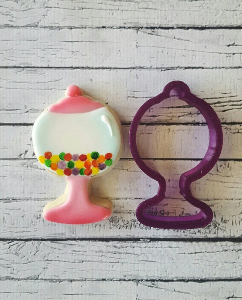 May include: A pink and white cookie cutter shaped like a gumball machine with colorful gumballs inside. The cookie cutter is next to a pink and white gumball machine cookie.