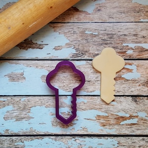 May include: Purple plastic key-shaped cookie cutter on a wooden background with a rolling pin and a beige plastic key-shaped cookie cutter.