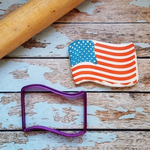 May include: A purple plastic cookie cutter shaped like a wavy rectangle, next to a red, white and blue American flag cookie on a wooden surface.