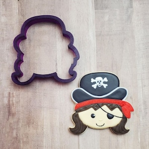 May include: A cookie cutter and a decorated sugar cookie in the shape of a pirate girl's head. The cookie has brown hair, a black eye patch, a red bandana, and a black pirate hat with a white skull and crossbones.