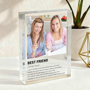 Puede incluir: Un bloque acrílico transparente con una foto de dos mujeres sonriendo. El bloque presenta el texto "BEST FRIEND" con una definición. Un portavelas geométrico dorado y una planta en maceta están en el fondo.