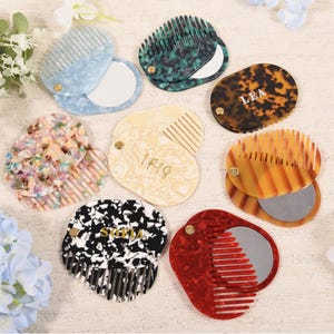 Puede incluir: Una variedad de peines compactos con espejos. Los peines presentan varios patrones y colores, incluyendo azul, verde, carey y rojo. Algunos peines tienen nombres como "LEA" e "IR19" impresos. Los peines están dispuestos sobre una superficie clara.