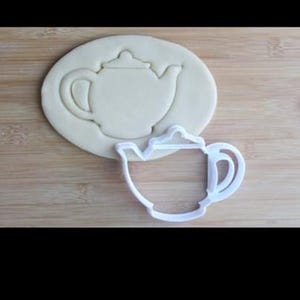 Peut inclure: Un emporte-pièce en plastique blanc en forme de théière à côté d'une découpe de théière dans de la pâte. Le découpeur a une anse et un bec, et la découpe de la pâte montre le contour de la théière. Sur une surface en bois brun clair.