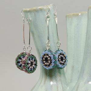 Peut inclure: Deux paires de boucles d'oreilles perlées avec des crochets d'oreilles argentés. Une paire présente un motif rond multicolore avec des perles vertes, roses et noires. L'autre paire a un motif perlé bleu, vert et noir.