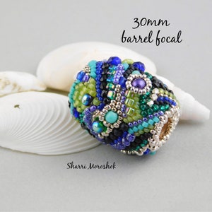 May include: A 30mm barrel focal bead with a colorful mosaic design. The bead is made with a variety of small beads in shades of blue, green, purple, and silver. The bead is sitting on a white seashell.