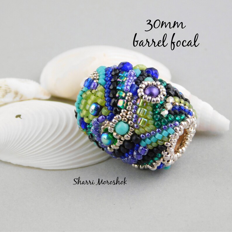 May include: A 30mm barrel focal bead with a colorful mosaic design. The bead is made with a variety of small beads in shades of blue, green, purple, and silver. The bead is sitting on a white seashell.