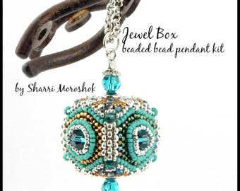 May include: A teal, gold, and silver beaded pendant kit with a silver chain. The pendant is shaped like a cube with a teardrop-shaped bead hanging from the bottom. The kit is called "Jewel Box beaded bead pendant kit" by Sharri Moroshok.